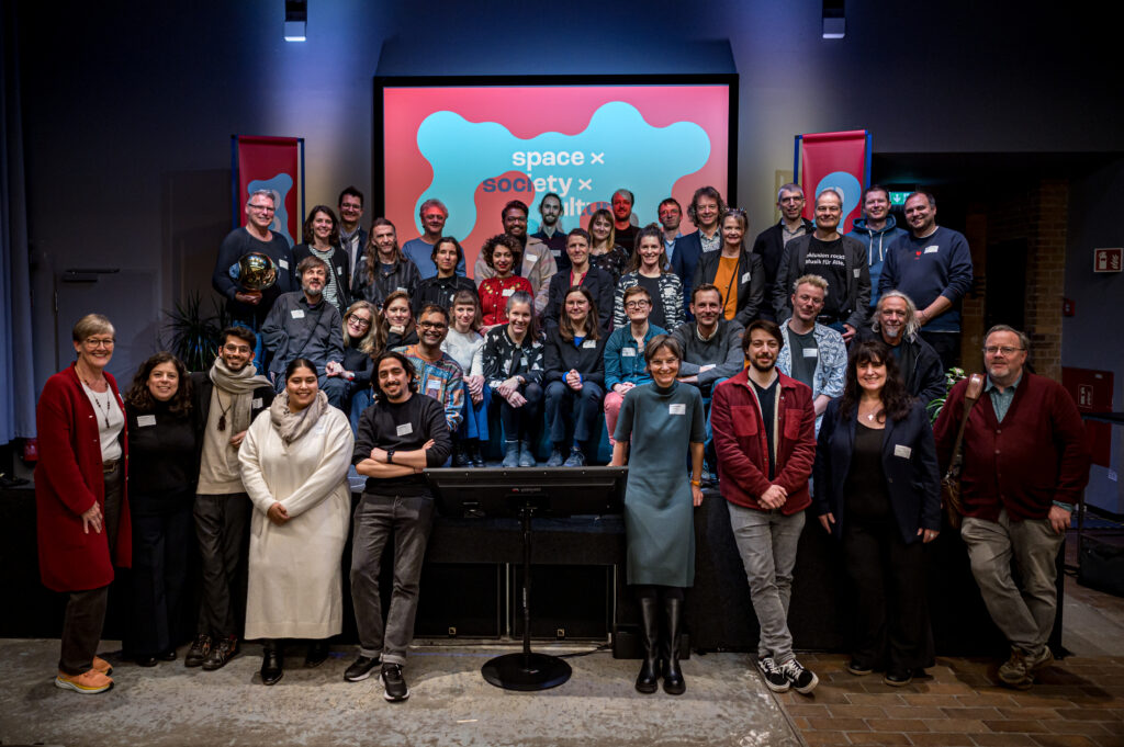 Gruppenfoto fon 38 Menschen auf und vor einer Bühne gruppiert, alle lächeln und tragen Namesnschilder, mit Hintergrund eine Leinwand mit Schrift "space x, society x"