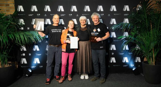 Buntfoto mit von rechts nach links: Peter, Claudia Roth, Jana und Thorsten. Jana, Peter und Thorsten tragen -handiclapped T-Shirts, alle stehen vor einer Fotowanbd mit dem Applaus Logo vielfach abgebildet. Jana hält eine Urkunde, Peter eine Plakette.