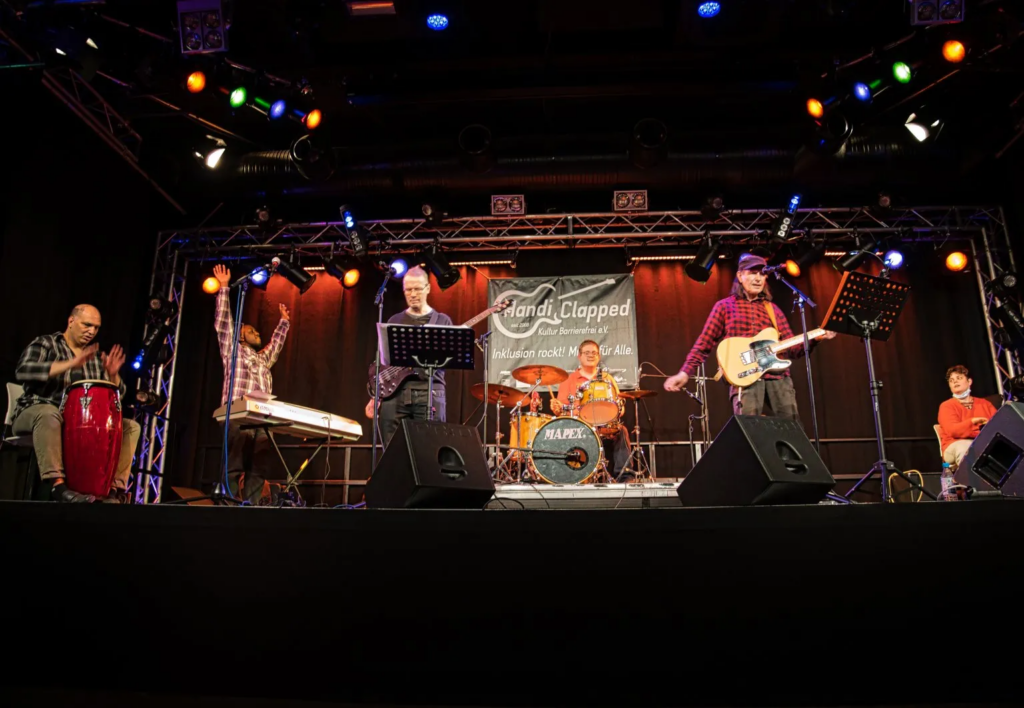 Die 6 Musikerinnen der Band auf der Bühne. Der Keyboarder links im Bild reckt die Arme nach oben.