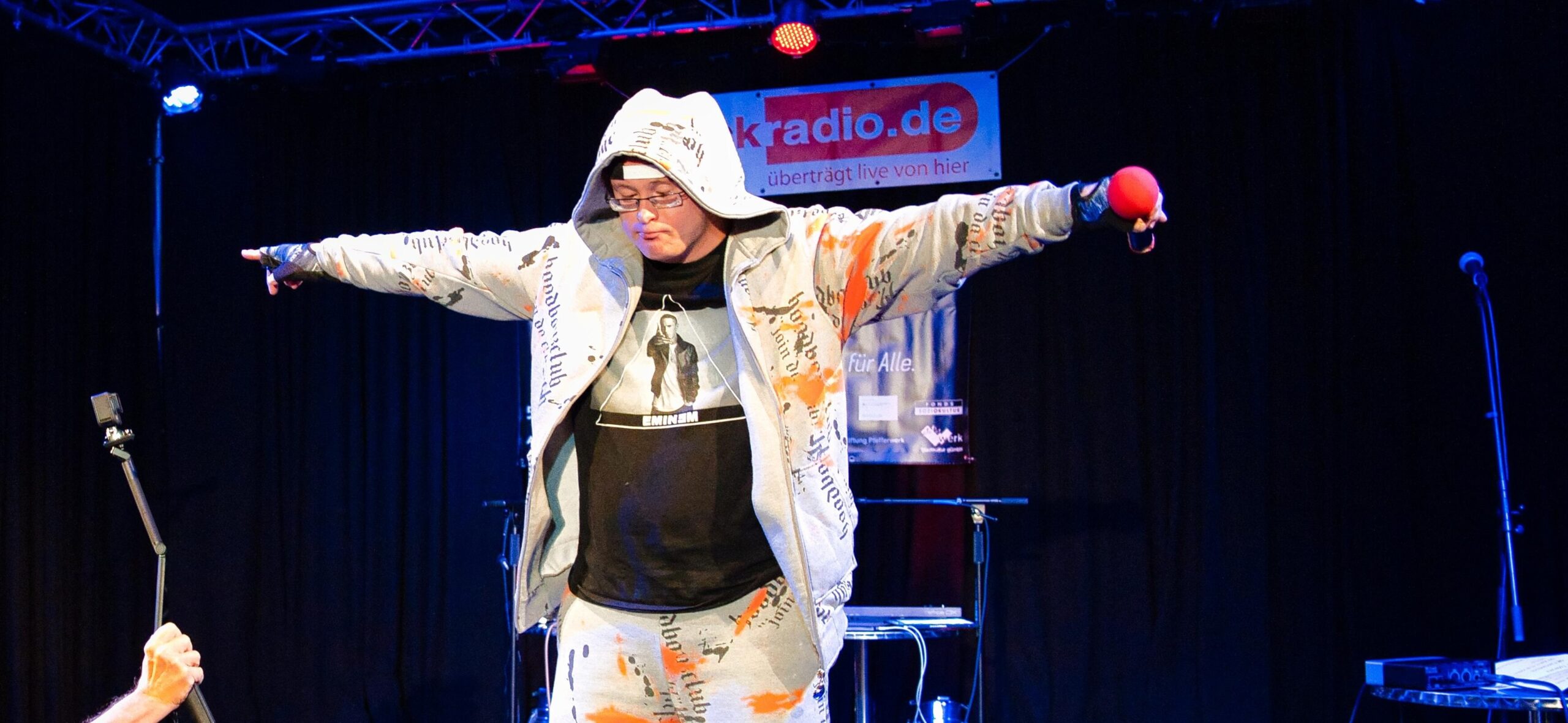 Buntfoto von einem jungen Mann in einem weißen Trainingsanzug udn schwarzen T-Shirt, der ein rute Mikrofon hält . Link im bild eine Kamera, die ihn filmt.
