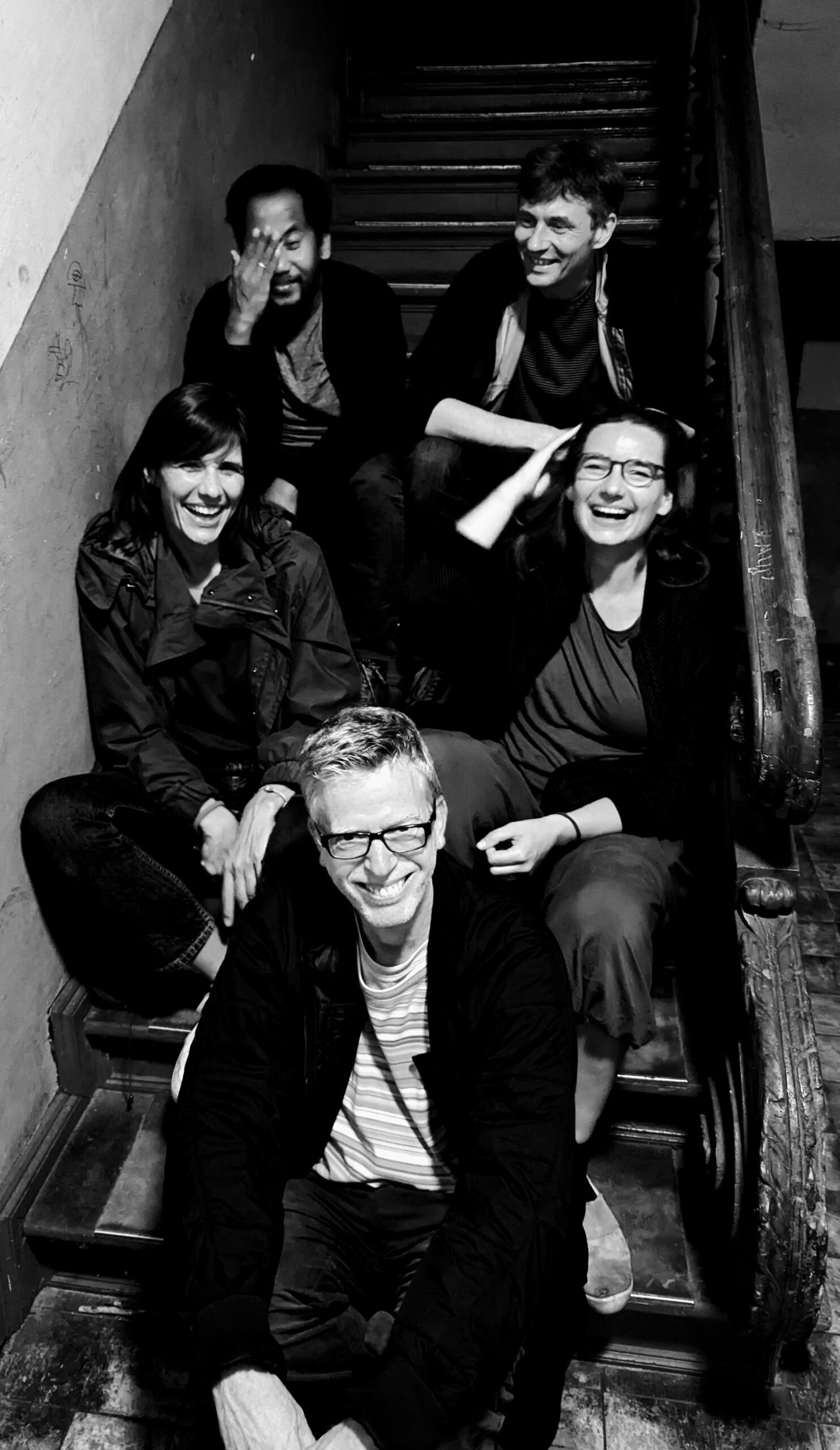 Ein schwarz weiß Foto von fünf gut gelaunten Menshen die in drei Rehien auf einer Treppe in einem alten Wohnhaus sitzen und in die Kamera lachen.