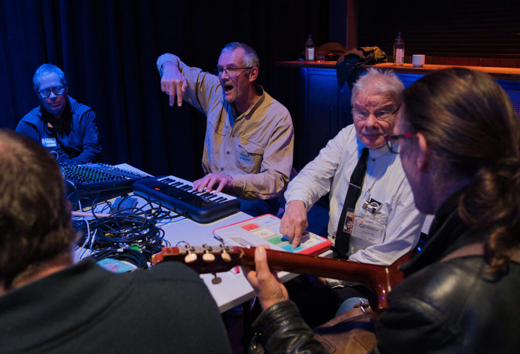 Buntfoto von einer Gruppe von fünf Personen, die um einen Tisch sitzen, auf dem ein Keyboard, ein Mischpult und andere Geräte stehen. Eine der Personen hält eine Gitarre.