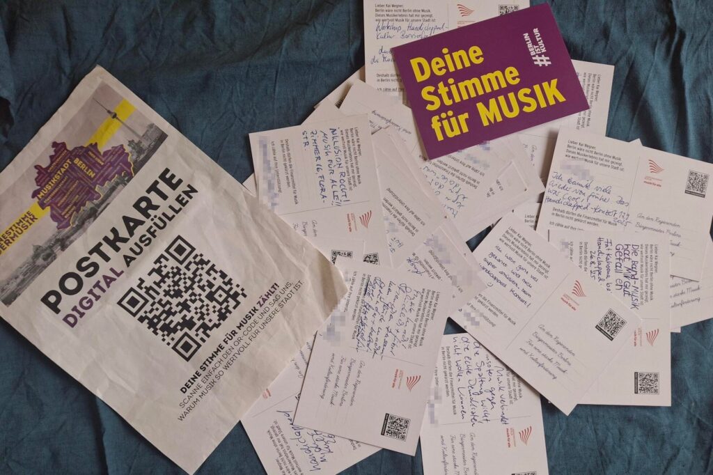 Ein Buntfoto von einem Haufen von ca 30 Postkarten, mit der Rückseite nach oben und die auf einem blauen Tuch ausgebreitet sind. Texte der Karten sind teilweise zu erkennen, die von Konzerten schwärmen. Unterschriften sind verpixelt. Eine Karte ist von Vorne zu sehen. Auf ihr steht: "Deine Stimme für Musik". Dazu ist ein Zetttle mit Barcode abgebildet, auf dem steht "Postkarte digital ausfüllen".