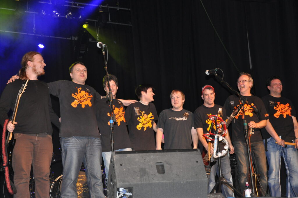 (Auf dem Bild sind 8 Musiker:innen in einer Reihe zu sehen. Wahrscheinlich am Ende ihres Konzertes, sie sehen glücklich und zufrieden aus.)