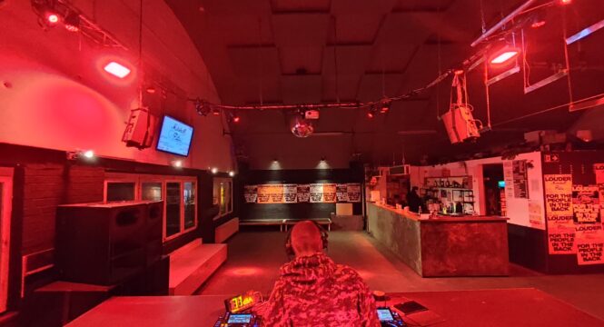 Buntfoto von einem Nachtclub, in rotes Licht getaucht. Im Vordergrund steht mit dem Rücken zur Kamera ein DJ und legt an zwei Plattenspieler und Technik auf, dahinter ein leerer Clubraum, geräumig mit Tanzfläche und langer Bar.