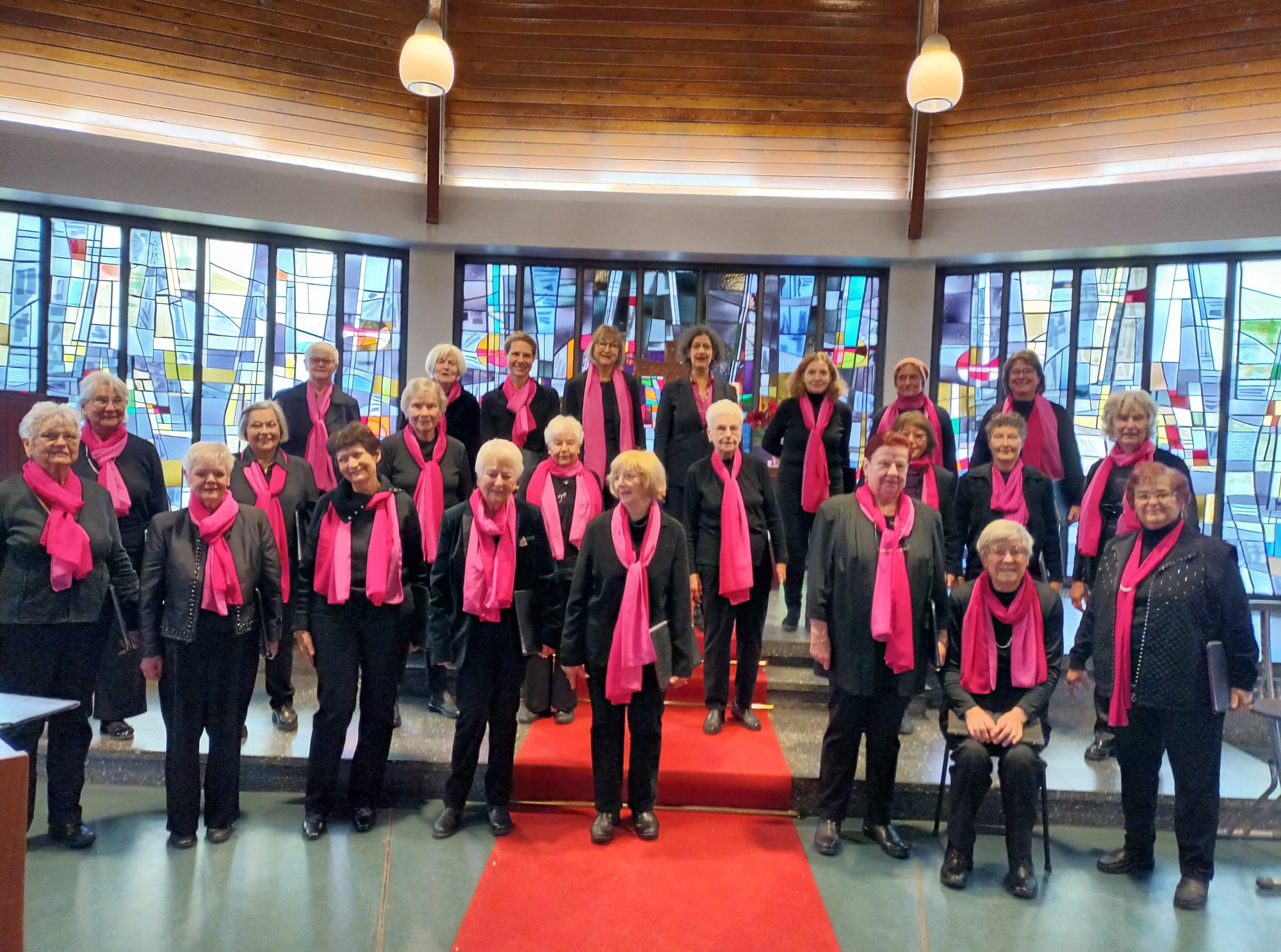 Buntfoto von 23 Damen in schwarzer Keidung und mit pinken Schals, vor einem riesiegen bunten Fenster. 