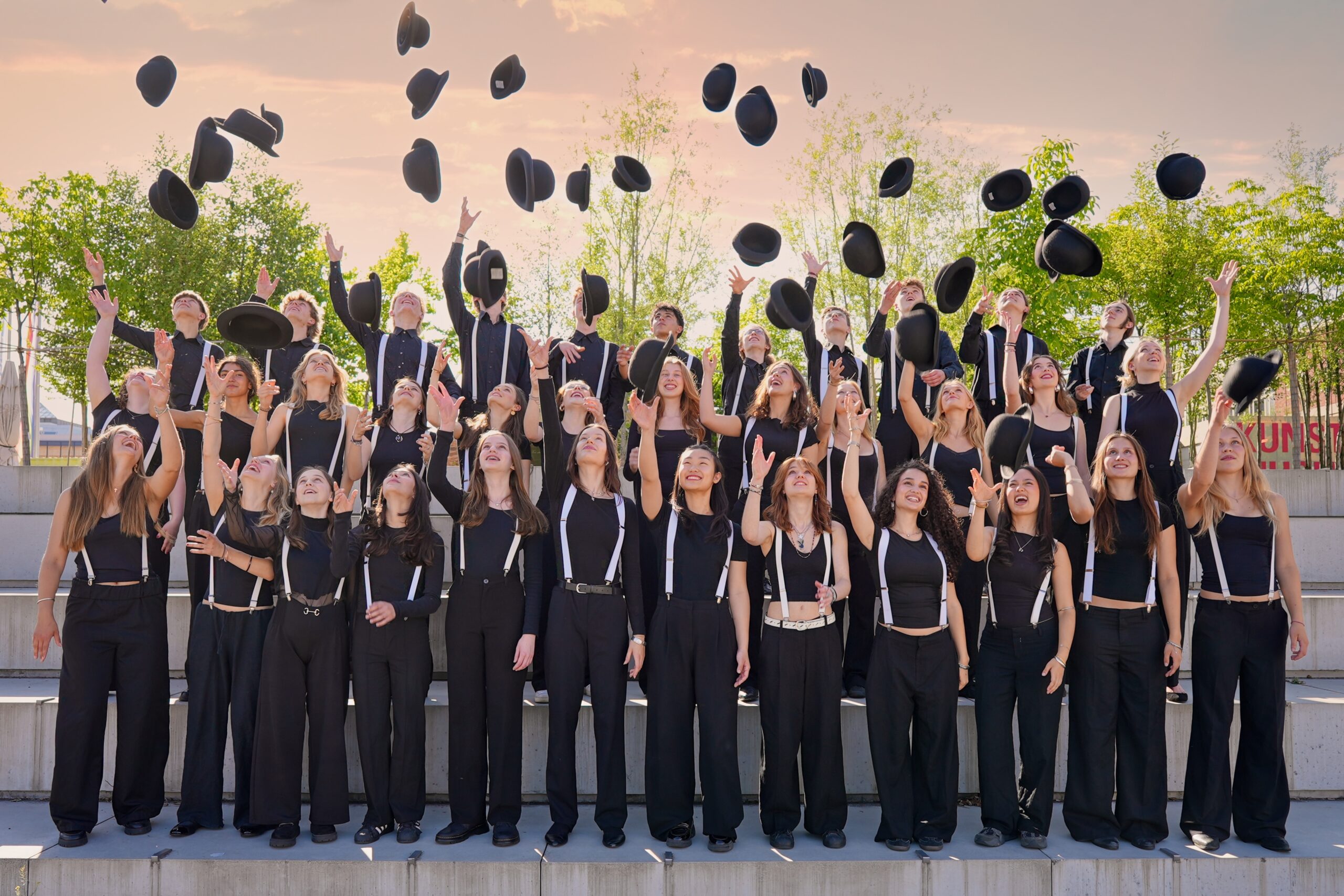 Buntfoto von 35 jugendlichen in schwarzer schicker kleidung in drei Reihen hinteereinander aufgestellt. Alle werfen einen schwarzen Hut in die Luft. Die Hüte schweben so auf dem Foto über der Gruppe.