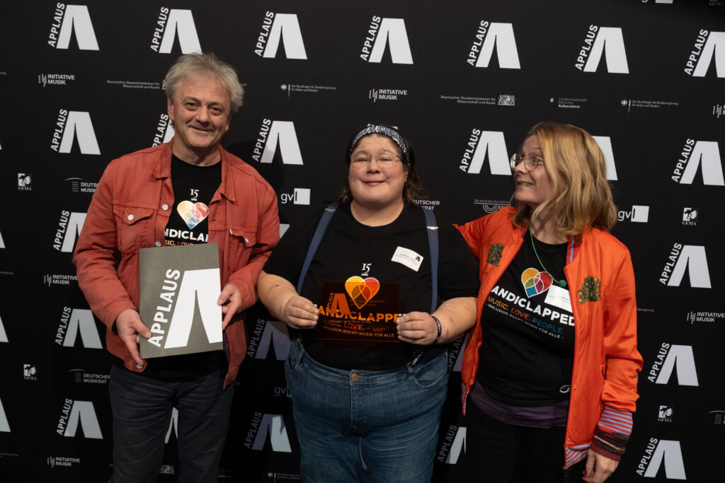 Jana, Jacqueline und Peter vor der Fotowand vom Applaus in München. In ihren Händen halten sie den Preis.
