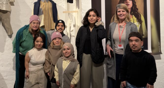 Eine Gruppe von Menschen, überwiegend Kleinwüchsige, schaut in die Kamera. Sie steht vor textilen Ausstellungsstücken und einem großen Plakat. Auf dem Plakat ist eine kleinwüchsige Frau in modischer Kleidung zu sehen.