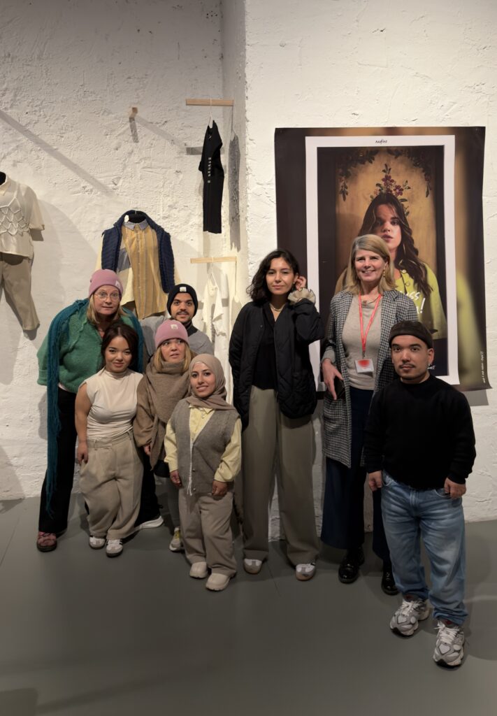 Eine Gruppe von Menschen, überwiegend Kleinwüchsige, schaut in die Kamera. Sie steht vor textilen Ausstellungsstücken und einem großen Plakat. Auf dem Plakat ist eine kleinwüchsige Frau in modischer Kleidung zu sehen.
