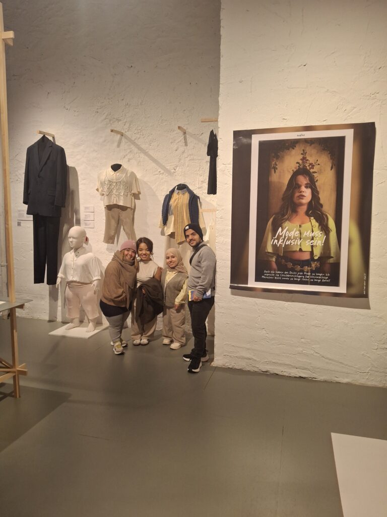 Vier kleinwüchsige Personen stehen, in die Kamera lächelnd, in einem Ausstellungsraum neben einer kleinwüchsigen Schaufensterpuppe. An der Wand hängt Mode für sehr große und sehr kleine Menschen. Rechts neben der Gruppe ist ein großes Plakat mit einem kleinwüchsigen Model. Der Slogan auf dem Plakat lautet: Mode muss inklusiv sein!
