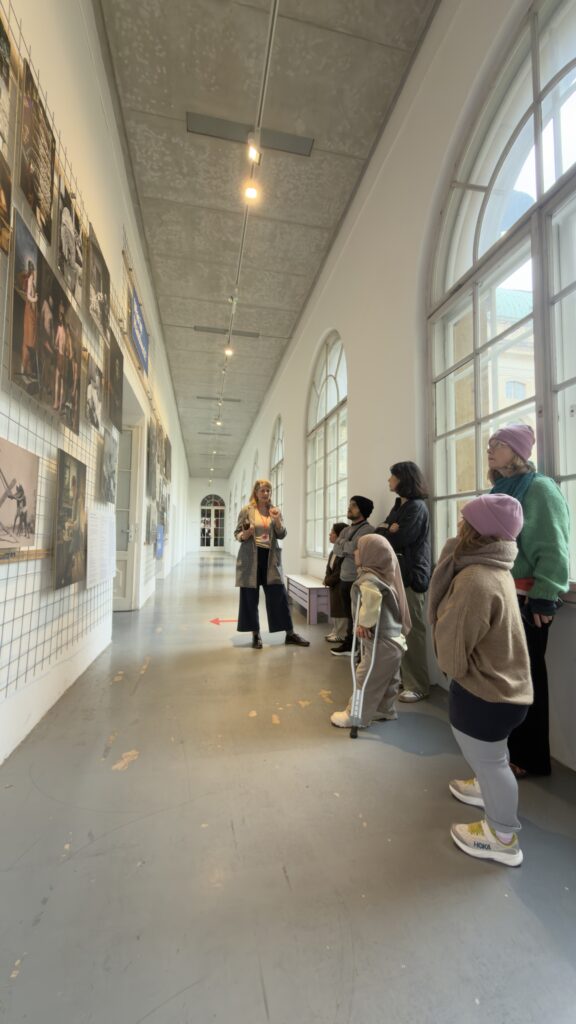 Eine Gruppe von Menschen schaut auf eine Wand mit ausgestellten Bildern zum Thema Handwerk. Eine Frau erklärt der Gruppe, was auf den Bildern dargestellt ist und was das mit dem Thema der Ausstellung Mythos Handwerk zu tun hat.