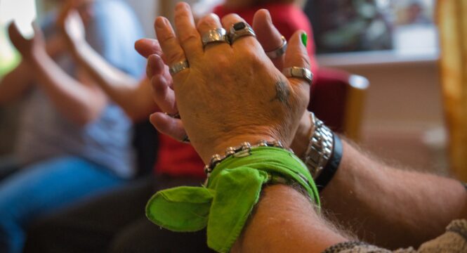 Nahaufnahme von klatschenden Händen mit mehreren silbernen Ringen und einem grünen Stoffband am Handgelenk; im unscharfen Hintergrund sitzen weitere Personen, die ebenfalls in die Hände klatschen