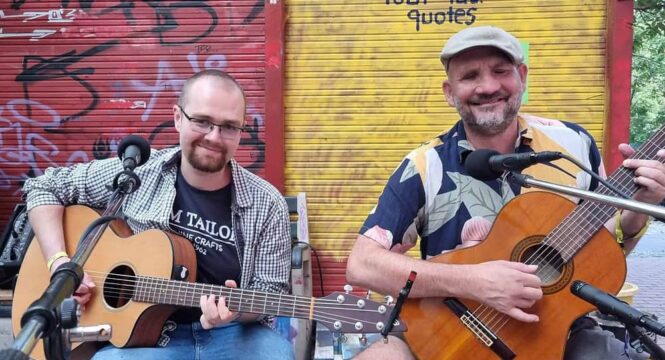 die 2 Musiker mit Gitarren sitzen vor einer rot-gelben Wand mit Graffities