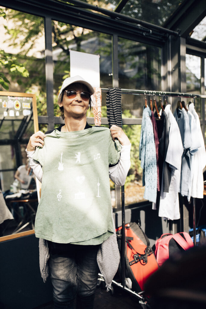 ein Frau mit dunkler Brille hält sich ein T-Shirt an und zeigt es in die Kamera. Es ist mit aufgebügelten Symbolen, wie Fernsehturm, Gitarre, Notenschlüssel versehen.