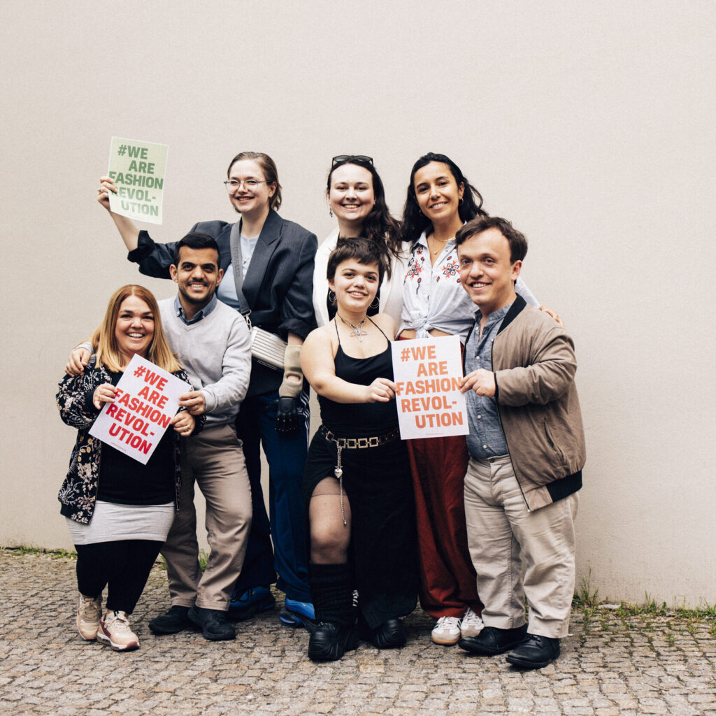 Buntfoto vom sieben Menschen als Gruppenfoto, lächelnd  halten Sie drei Zettel mit Schrift "#we are fashion revolution" hoch