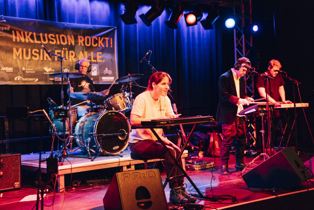 eine Band auf der Bühne, im Vordergrund eine Frau am Keyboard, links hinter ihr der Schlagzeuger. Rechts im Bild ein weiterer Keyboarder und ein Mann mit Kopfhörern an E-Drums. Blau/rotes Bühnenlicht