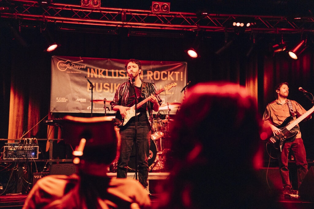 eine Band auf der Bühne, man sieht 2 Gitarristen in rotem Bühnenlicht, einige Köpfe vom Publikum von hinten im Vordergrund