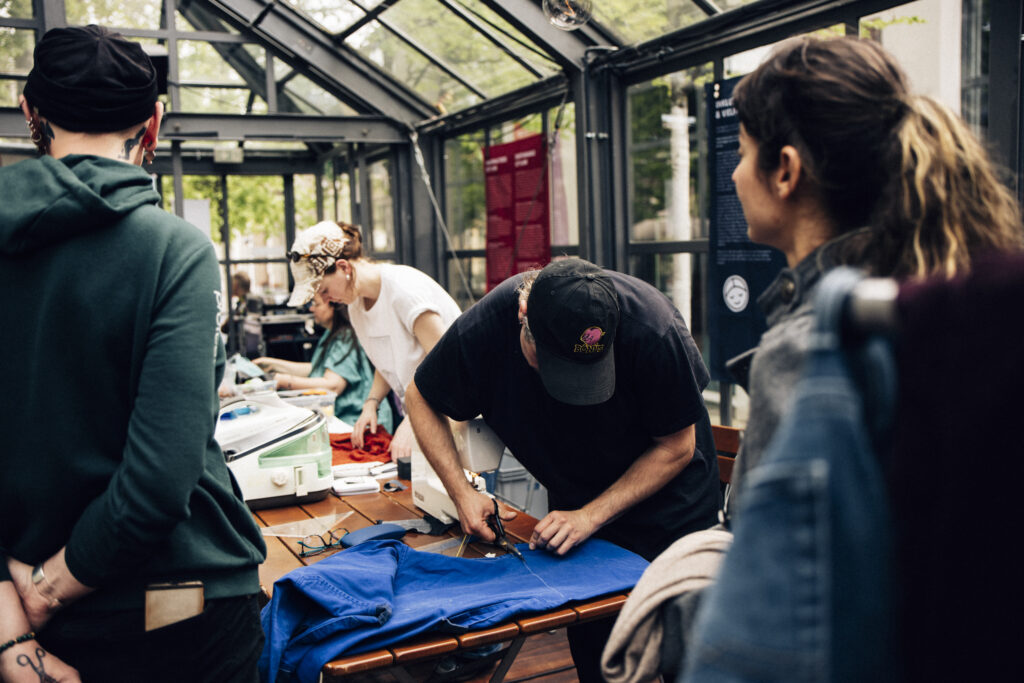 3 Personen werkeln an einem Tisch mit Kleidung, eine schneidet Stoff, eine andere ist an einer Maschine zum Bedrucken ... im Vordergrund 2 Personen, die zuschauen
