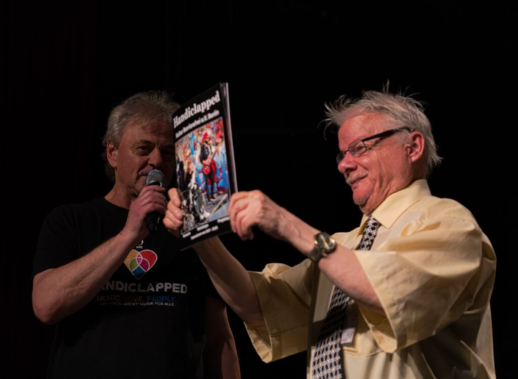 Achim und Peter auf der Bühne. Achim hält einen Handiclapped Fotoband hoch. 