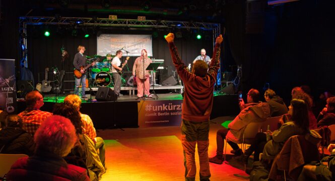 Buntfoto von einer Bühne (innen), darauf eine Band mit sechs Mitgliedern und Instrumenten, davor Publikum auf Stühlen sitzend, in der Ganggasse steht ein Mann und reckt die Arme zur Decke. Das Foto ist von hinter dem Publikum aufgenommen.