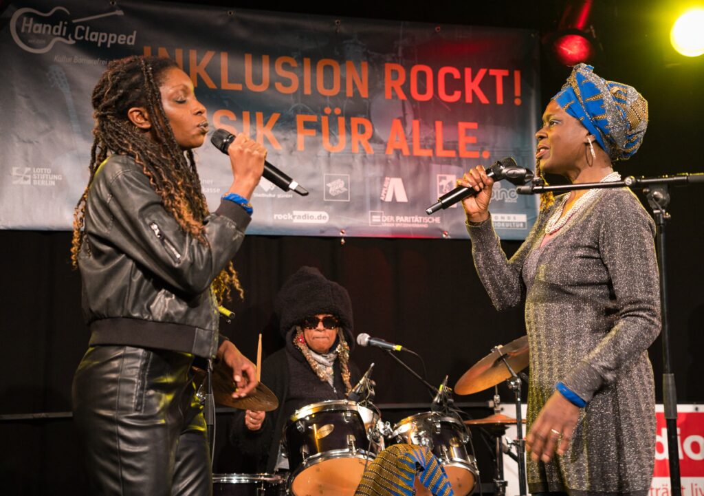 Buntfoto von zwei Frauen , die sich auf einer Bühne gegenüber stehen und beide in je ein Mikrofon singen. Im Hintergrund eine Schlagzeugerin in Aktion.