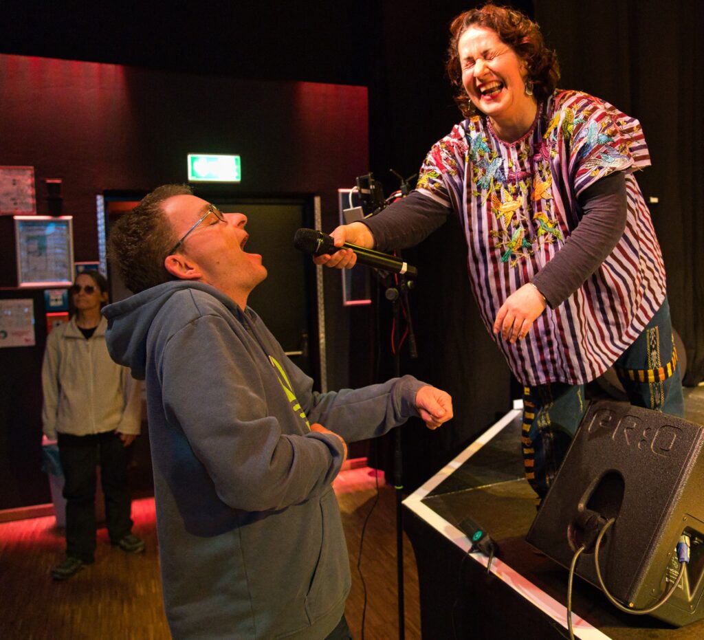 Buntfoto von einer lachenden Frau die von der Bühne herab ein Mikro an einen jungen Mann im Publikum hält der glücklich in dieses Mikrofon singt. 