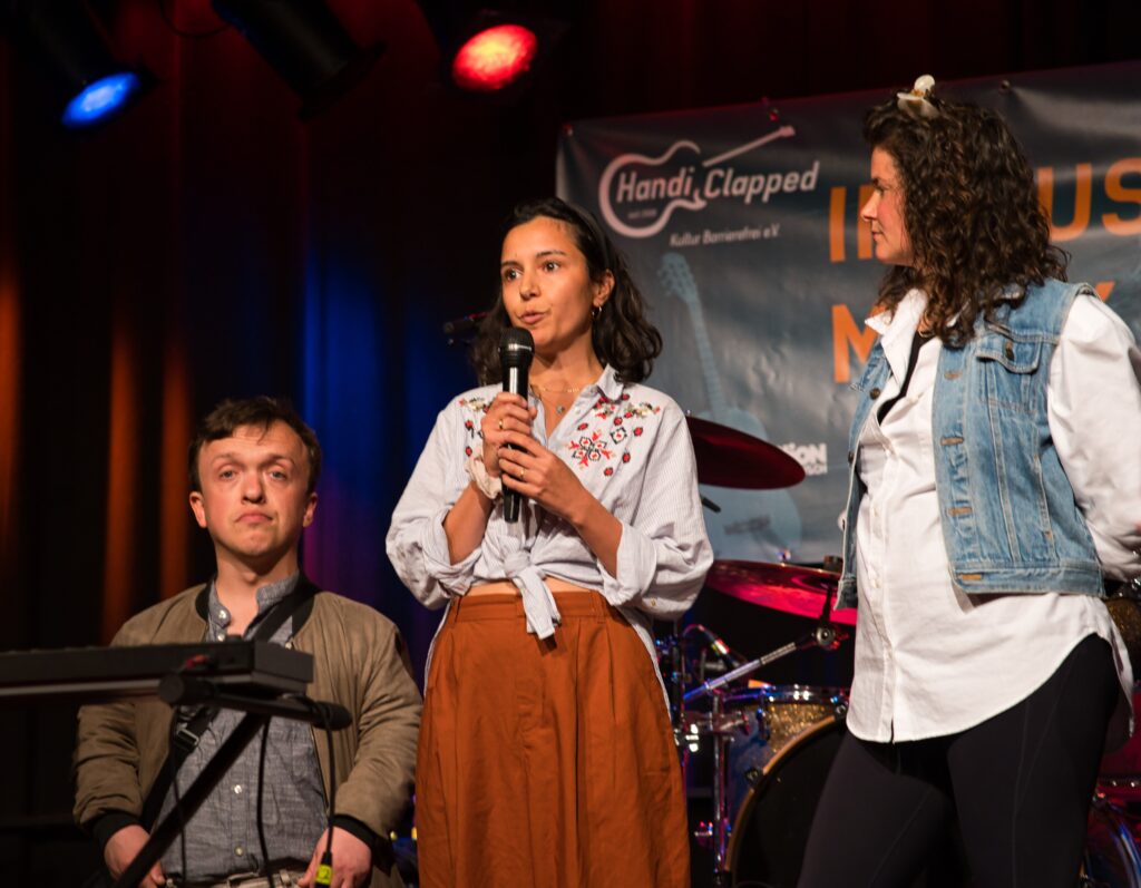 Buntfoto von drei Menschen auf einer Bühne, recht ein kleinwüchsiger Mann, in der Mitte eine junge Frau die in ein Mikrofon spricht, links eine weitere junge Frau.