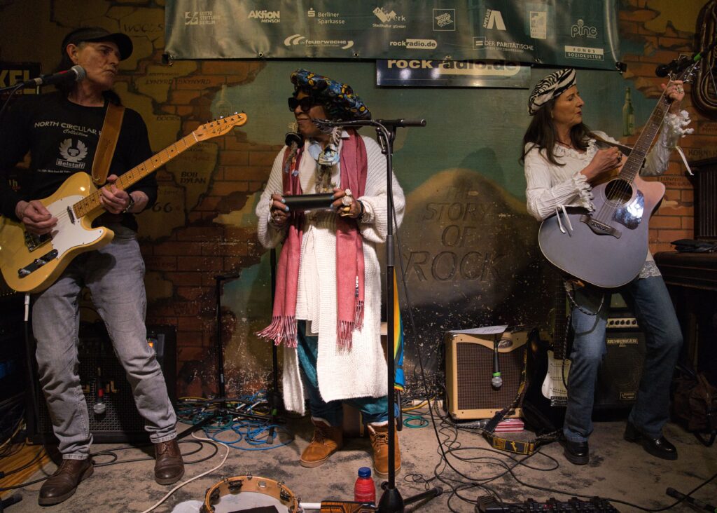Behind the Curtains beim Konzert, rechts ein Gitarrist, in der Mitte die Sängerin mit einer Rassel, rechts ein Gitarristin 
