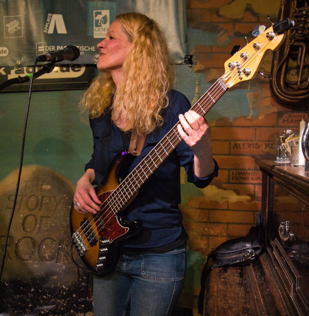 eine blonde, langhaarige Bassistin mit Instrument am Mikrophon