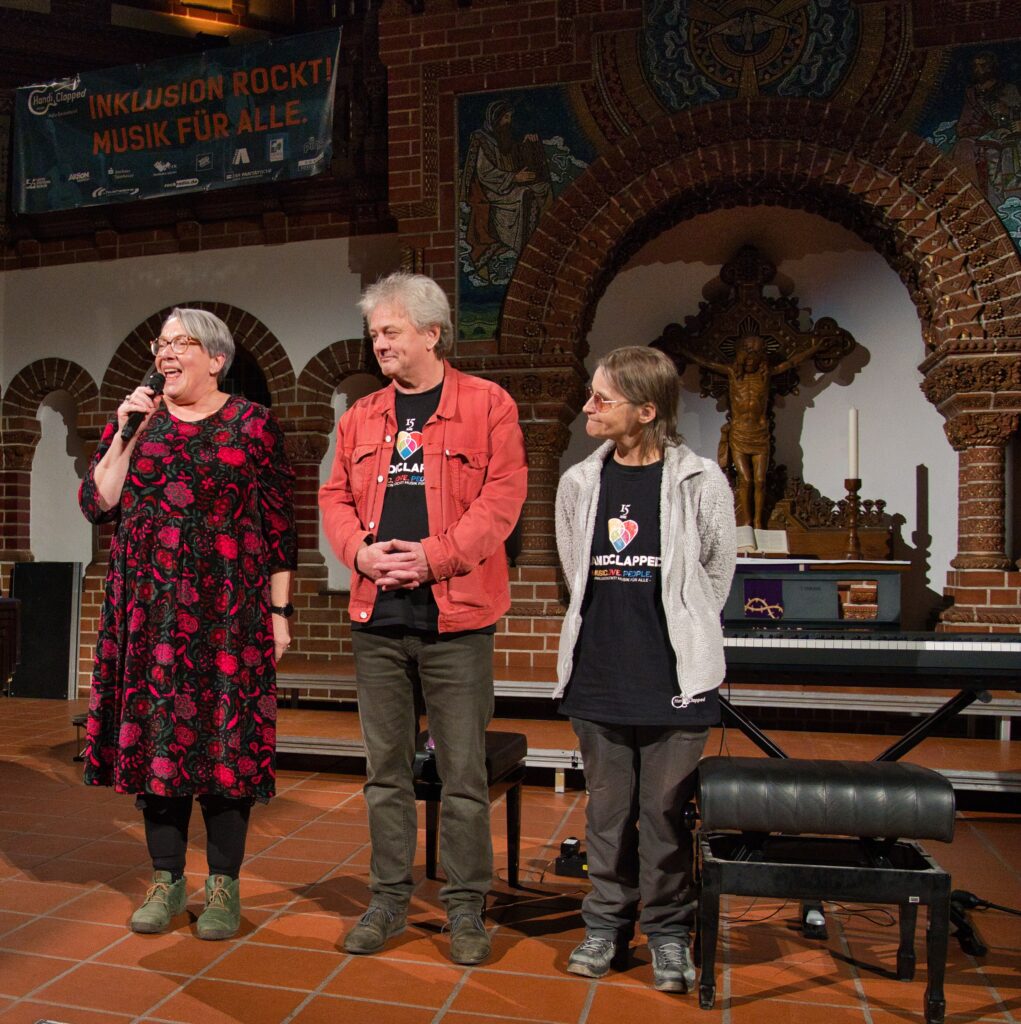 Drei Personen stehen in einer Kirche, ein hat ein rot schwarzes Kleid an und spricht in ein Mikrofon welches sie Held, die anderen beiden tragen T-Shirts mit Schrift "Handiclapped"