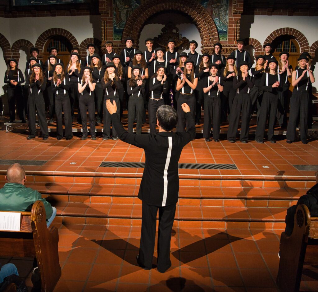 Buntfoto von einem Chor aus ca 30 jungen Personen. Alle stehen, tragen schwarze lange Hosen, schwarze Oberteile, weiße Hosenträger und schwarze Hüte (Melonen). Sie stehen auf der Bühne in einer Kirche. Davor, mit dem Rücken zur Kamera eine Dirigentin beim Dirigieren, ebenfalls in schwarz gekleidet.

