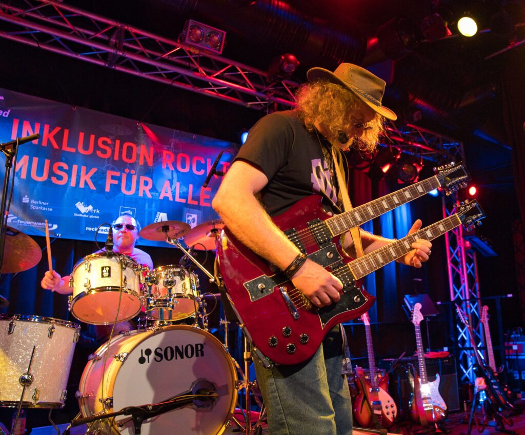 ein Gitarrist mit Hut und langen lockigen Haaren an einer doppelhalsigen roten Gitarre. links hinter ihm der Schlagzeuger mit dunkler Brille