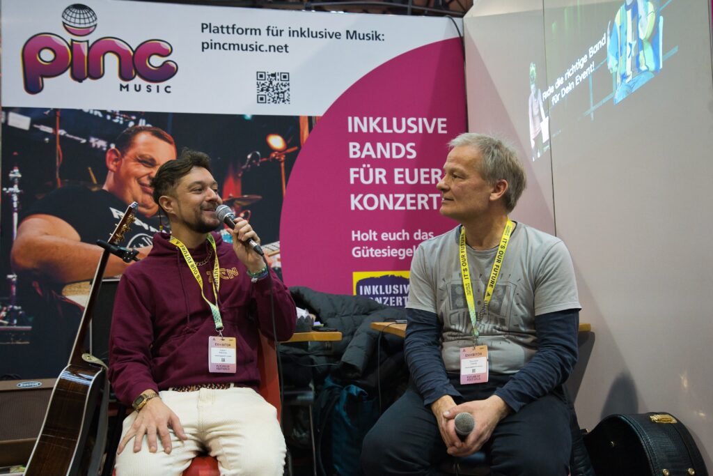 Buntfoto von einem Messestand an dem ein Mann einen anderen mit Mikrofon interviewt. Beide sitzen. Im Hintergrund ein Poster mit Foto eines Gitarristen und Pinc Music Logo und Schrift "Inklusive Bands für Euer Konzert"
