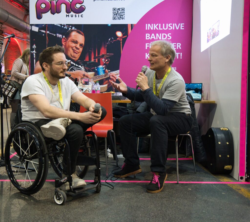 Buntfoto von einem Messestand an dem ein Mann einen anderen mit Mikrofon interviewt. Beide sitzen, davon einer in einem Rollstuhl. Im Hintergrund ein Poster mit Foto eines Gitarristen und Pinc Music Logo. 