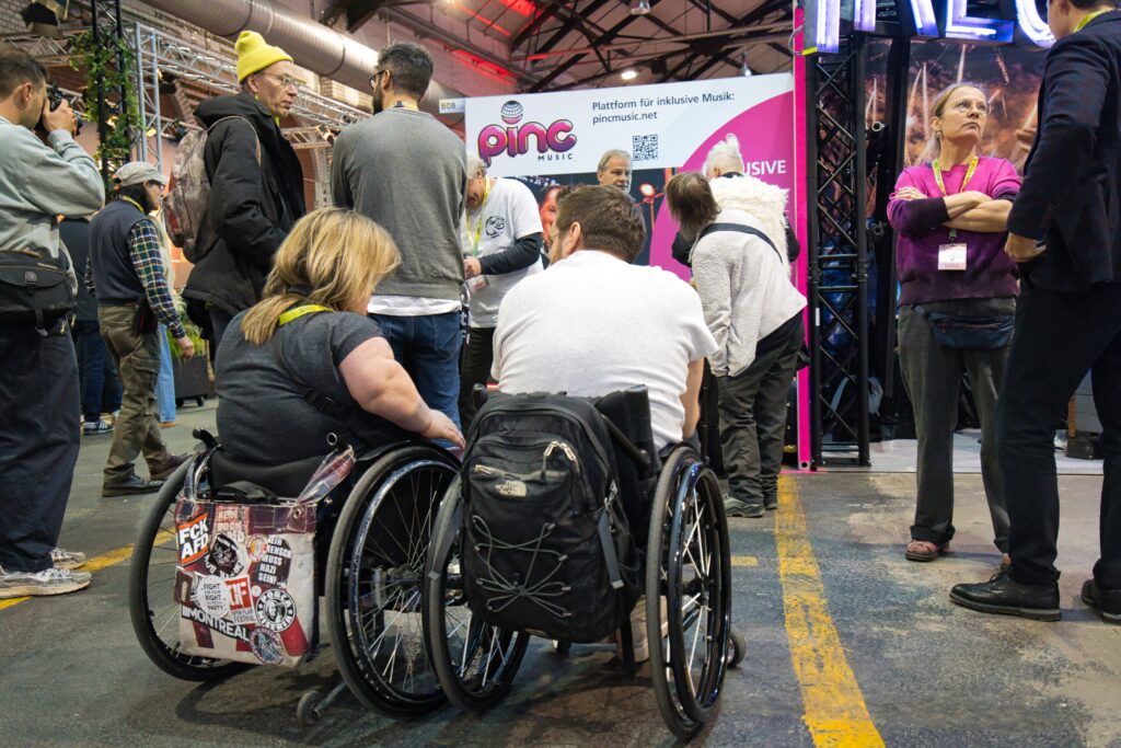 Buntfoto von einem Messestand, mit großem Pinc Music Logo. Davor Publikum stehen, zwei sitzen in Rollstühlen. 
