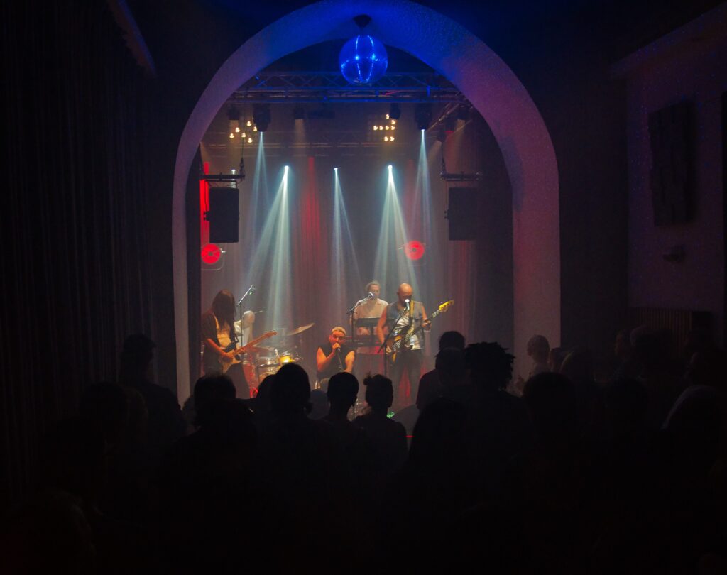 Buntfoto von einer Band auf einer kleinen Bühne, mit E-Gitarre, E-Bass, Keyboard und Schlagzeug, davor Publikum, darüber eine Diskokulgel. Lichtshow. Der Sänger singt in ein Mikro.
