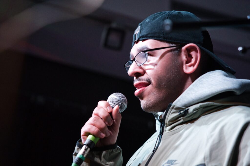 Nahaufnahme von einem  jungen Mann mit umgedrehter Schirmmütze und mit Brille. Er singt in ein Mikro