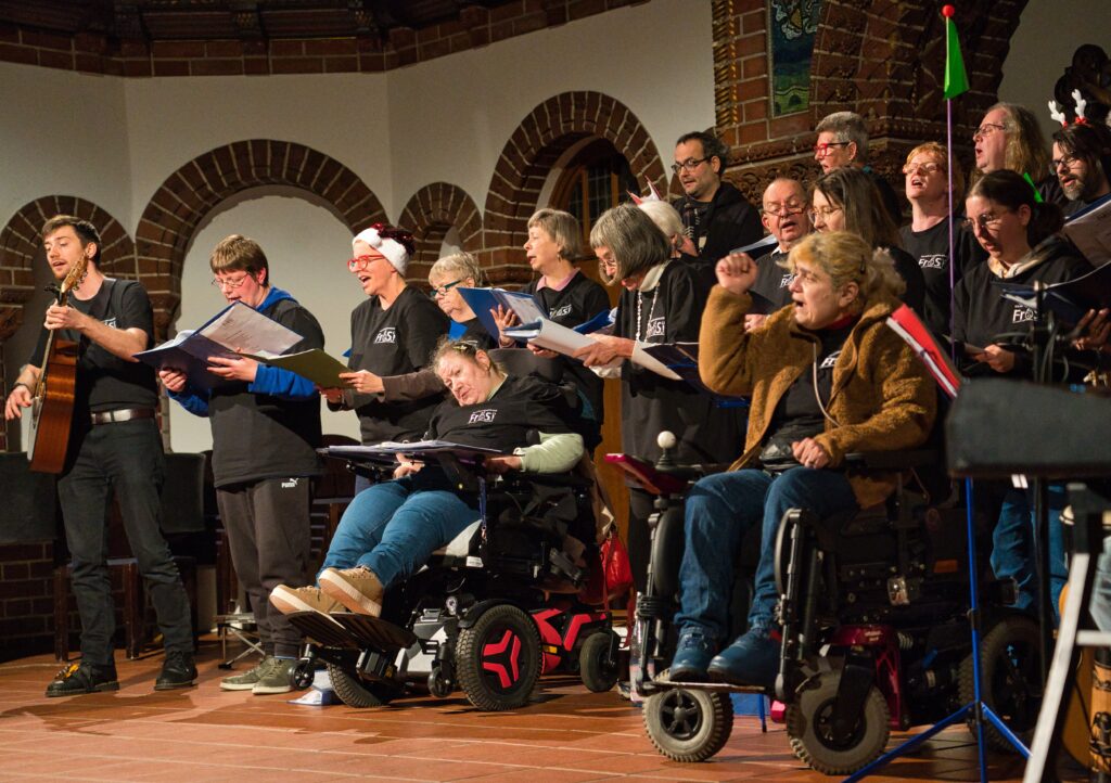 Ein Chor aus ca 20 Personen der in einer Kirche singt. Ein Mann spielt Gitarre, zwei Frauen sitzen im Rollstuhl. 