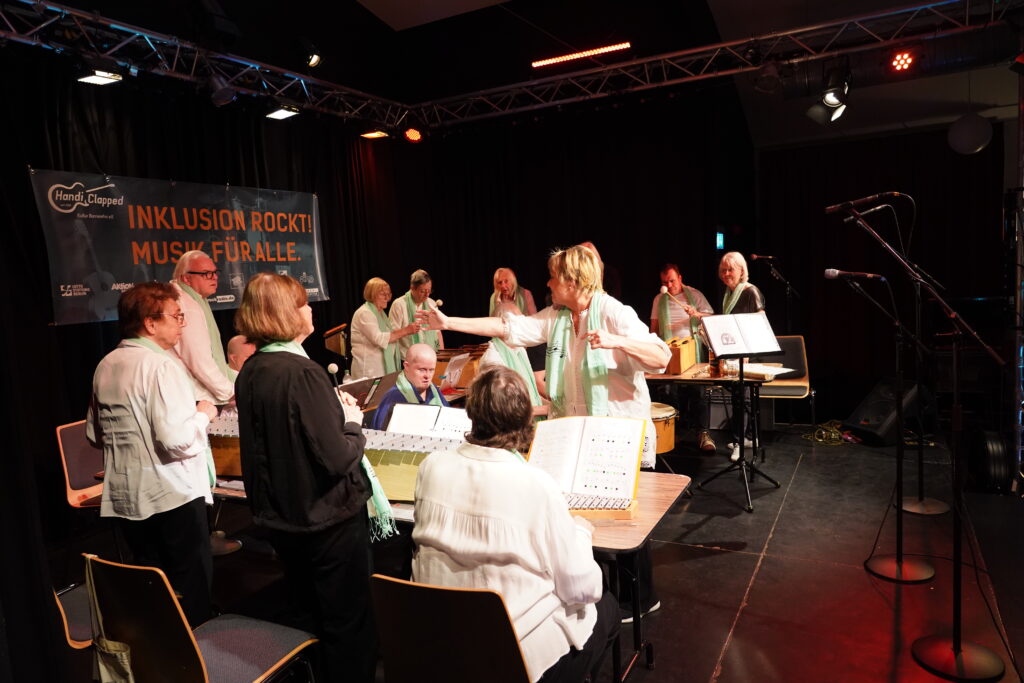 Elf Menschen auf einer Bühne, mit Glockenspielen und Trommeln. Eine Frau dirigiert.