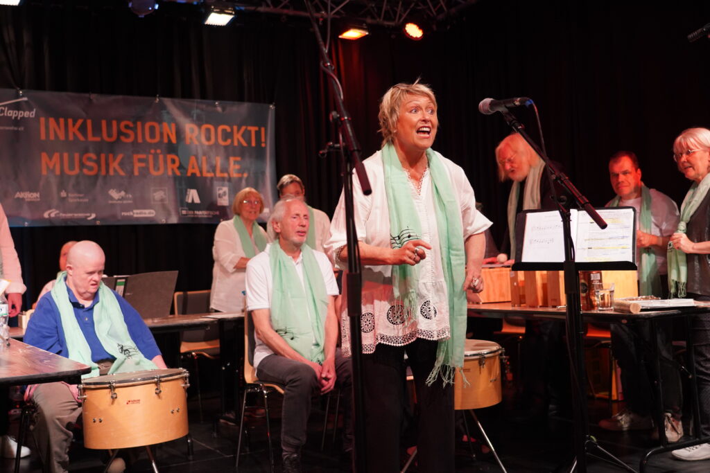 Neun Menschen auf einer Bühne, mit Glockenspielen und Trommeln. Eine Frau singt. Im Hintergrund Plakat mit Schrift "Inklusion rockt! Musik für Alle."