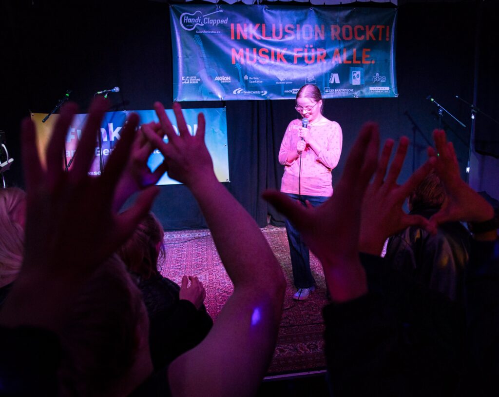 Eine Jugendliche singt auf einer Bühne in ein Mikrofon, im Vordergrund erhobene Hände vom Publikum