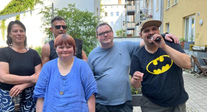 Buntes Gruppenfoto von fünf Menschen die auf der straße nebeneinander stehen und in die Kamera gucken. Eine trägt ein Batmah T-Shirt und singt in ein Mikro