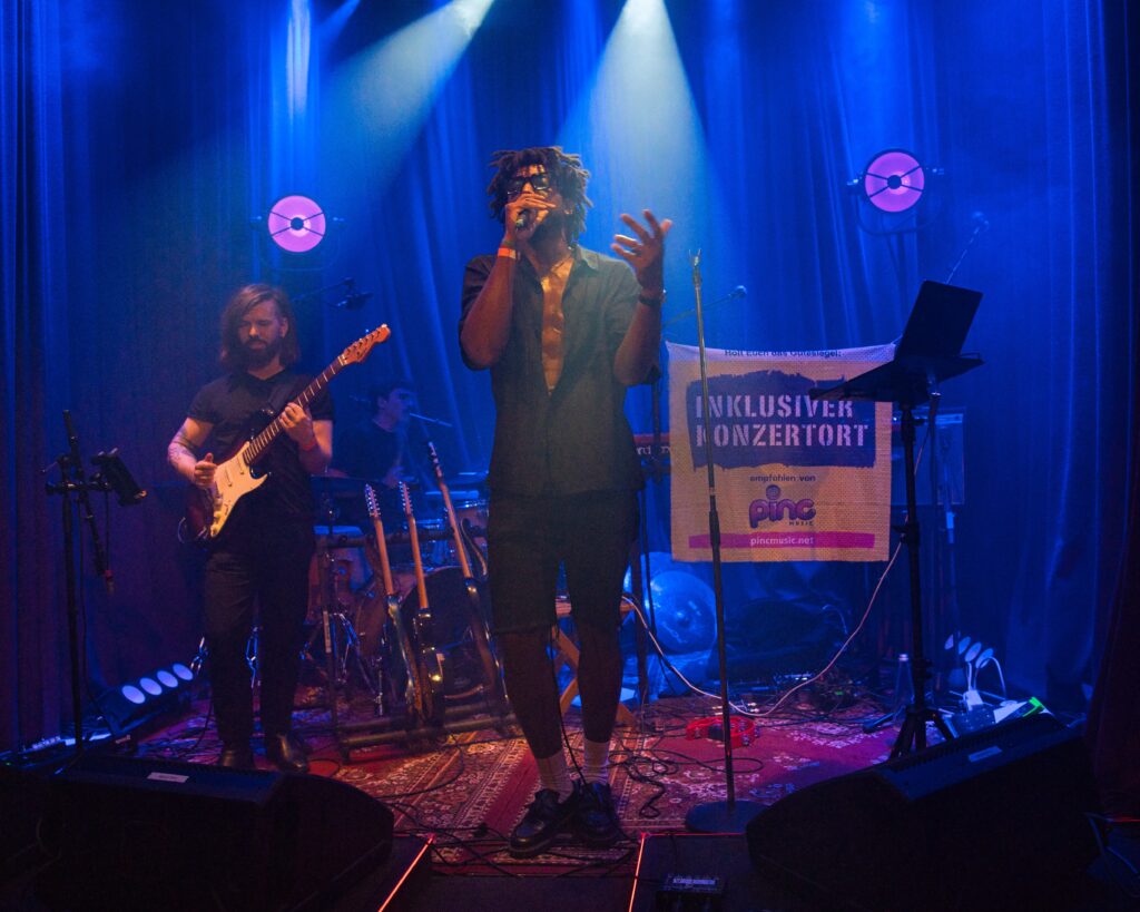 Drei Musiker beim Auftritt auf einer kleinen Bühne mit Lichtshow, der Sänger singt in ein Mikro dass er in der Hand hält. Einer spiel SChlagzeug, einer E-Gitarre, dazu ein Banner auf dem steht: "Inklusiver Musikort"