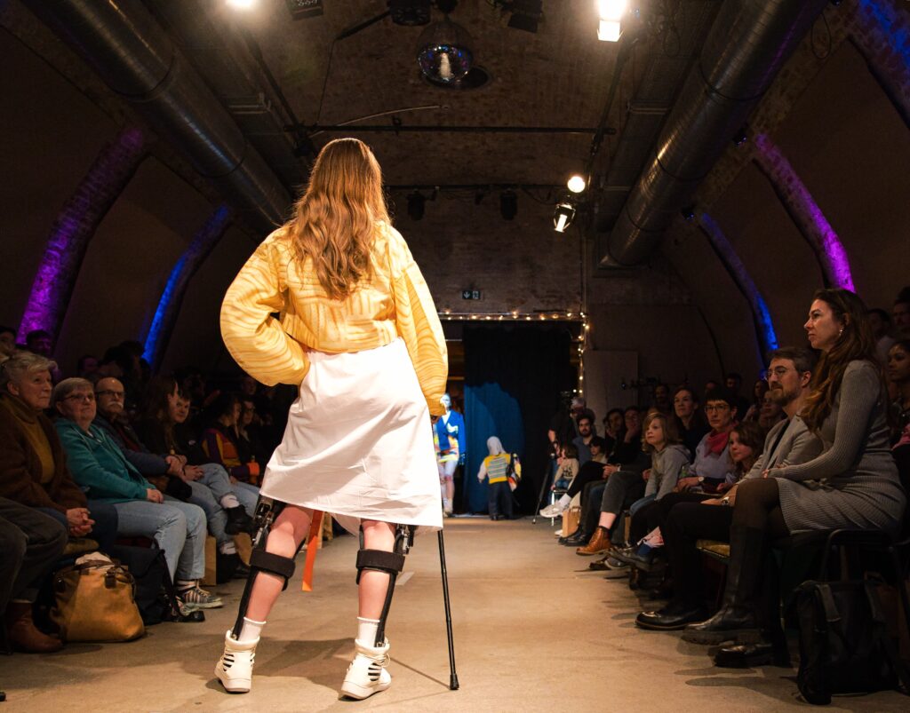 Buntfoto von einem Laufsteg auf einer Modenschau. Mit dem Rücken zur Kamera darauf eine Frau mit langen Haaren, gelbem Hemd und weißem Rock, Gehstock und Beinstützen. Rechts und links Publikum.