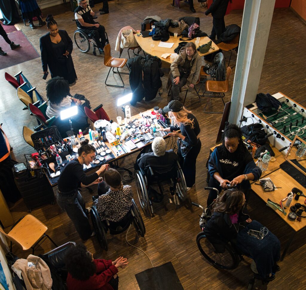 Backstage einer Modenschau von oben fototografiert, Schminktische an denen 3 junge Frauen in Rollies geschminkt und frisiert werden