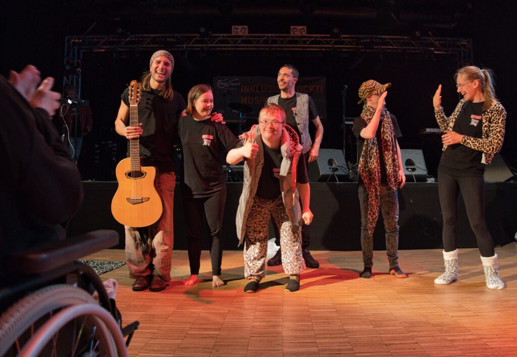 Buntfoto von sechs jungen Menschen, die nach einer Tanz-Performance glücklich teils ins Publikum gucken, zwei klatschen sich untereinander ab. 