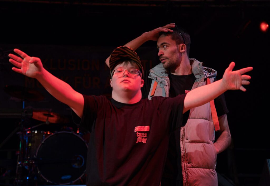 Buntfoto von zwei jungen Männern. Sie stehen hintereinander. Der vordere breitet die Arme aus und guckt ins Publikum, der hintere guckt zur Seite. 