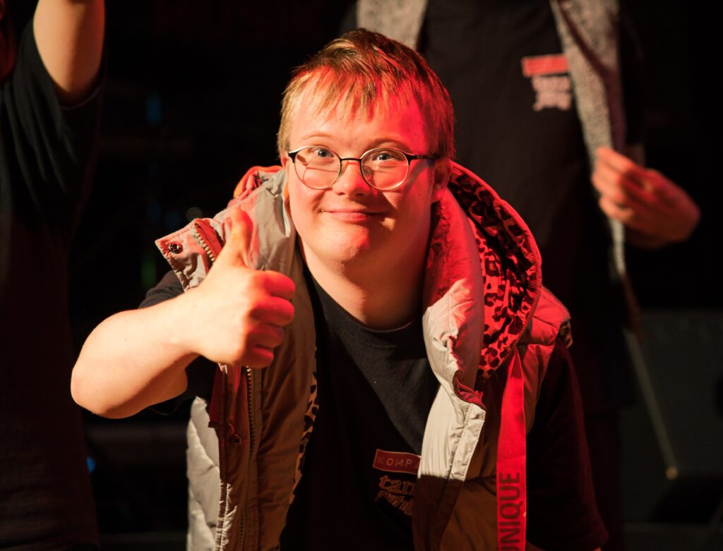 Buntfoto von einem jungen Mann der in die Kamera guckt und mit der Hand ein Daumen hoch gibt.