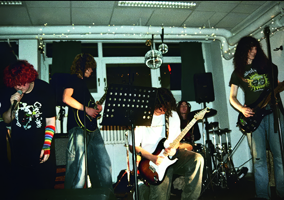 Buntfoto von einer Punk-Rock-Band, die in einem Keller auftritt mit Schlagzeug, E-Bass, E-Gitarre und langen Haaren.
